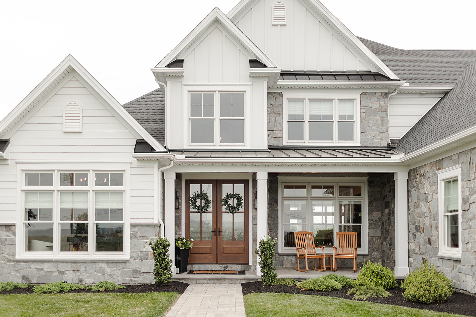 Modern two-story home exterior featuring white board-and-batten siding, stone facade accents, dark wood front doors, and a covered porch.